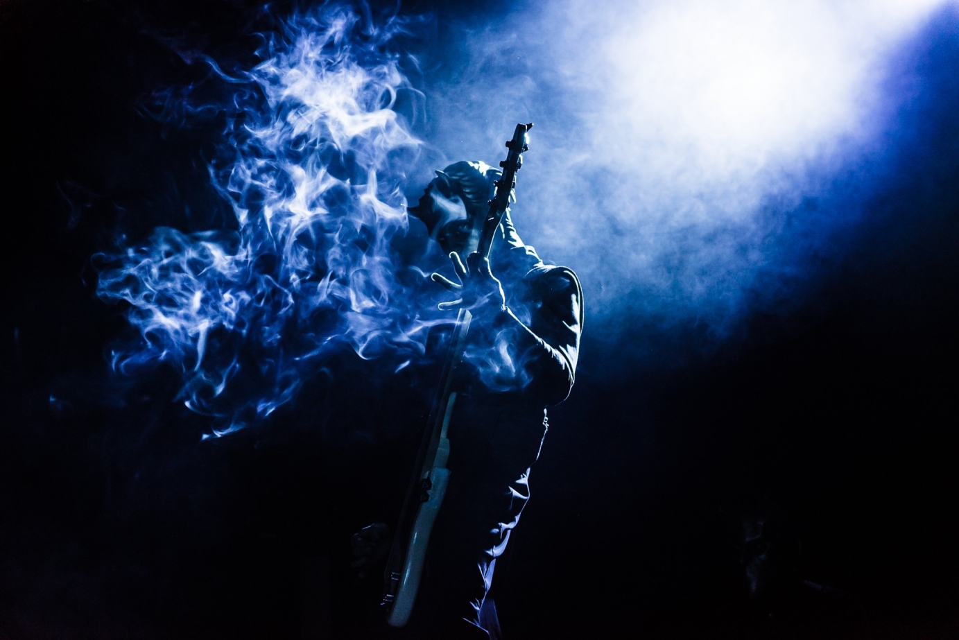 Guitarist from the band ghost, wearing a mask, smoke billowing out of the guitar, highlights and shadows brining an emotive feel to the darkness